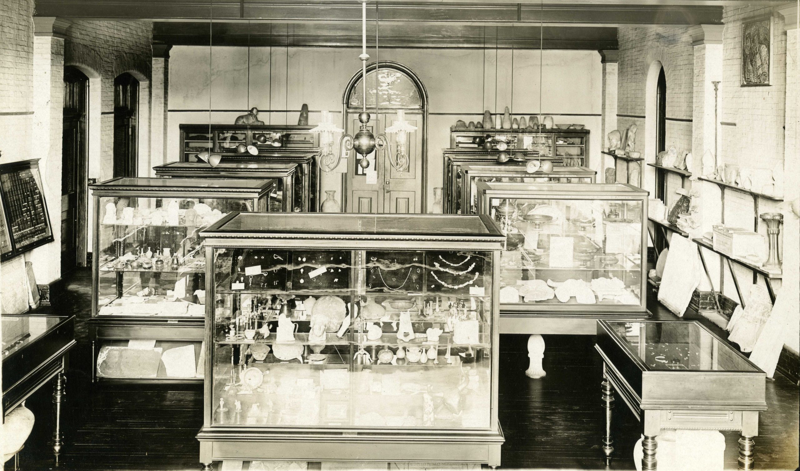 Historic photograph of the Johns Hopkins Archaeological Museum gallery in McCoy Hall, circa 1915. The image shows a well-lit room with glass display cases filled with ancient artifacts, including pottery, jewelry, and tools. Artifacts also line shelves mounted on the brick walls. A wooden door and arched window are visible at the far end of the room and a chandelier hangs from the ceiling.