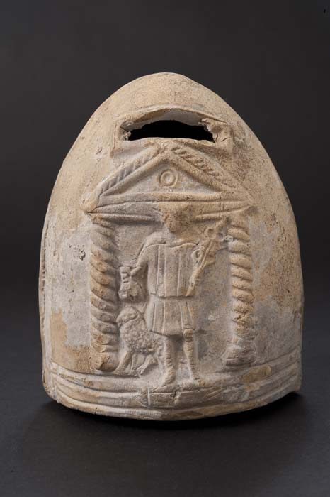 Photograph of an ancient Roman ceramic bank. The bank is roughly conical with a framed, rectangular hole in one side. Relief decoration on one side features two spiral columns and a triangular pediment, a standing figure in a knee-length tunic, and a sheep. The figure holds a small pouch in his right hand and a short staff with curling ends, perhaps a cadeuceus, in his left.