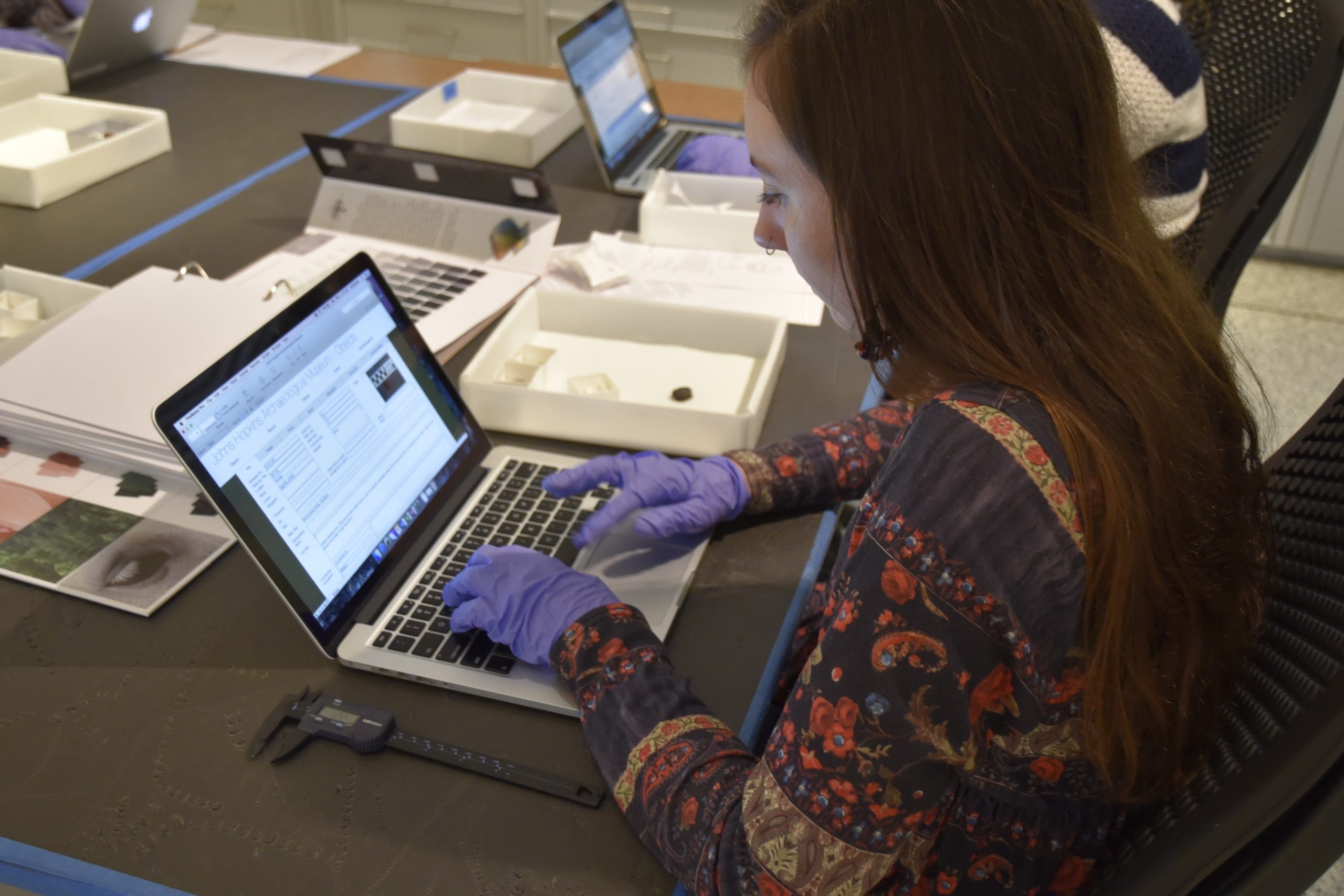 Photo of a woman with long brown hair wearing a floral patterned shirt sitting at a table in a museum typing on a laptop and working with an ancient Egyptian scarab.