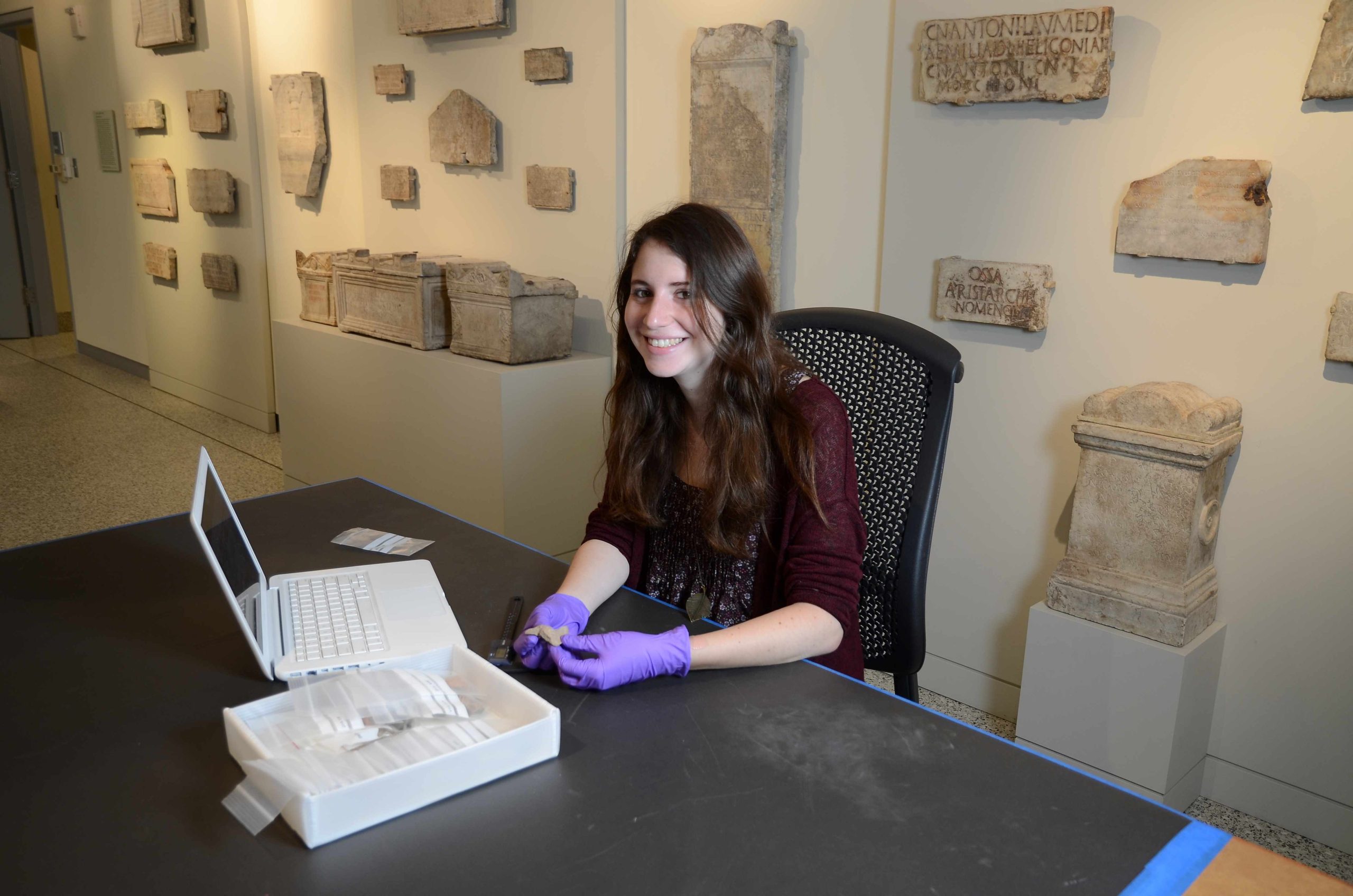 Photo of a woman with long brown hair sitting at table in a museum holding a vessel sherd in front of a laptop.