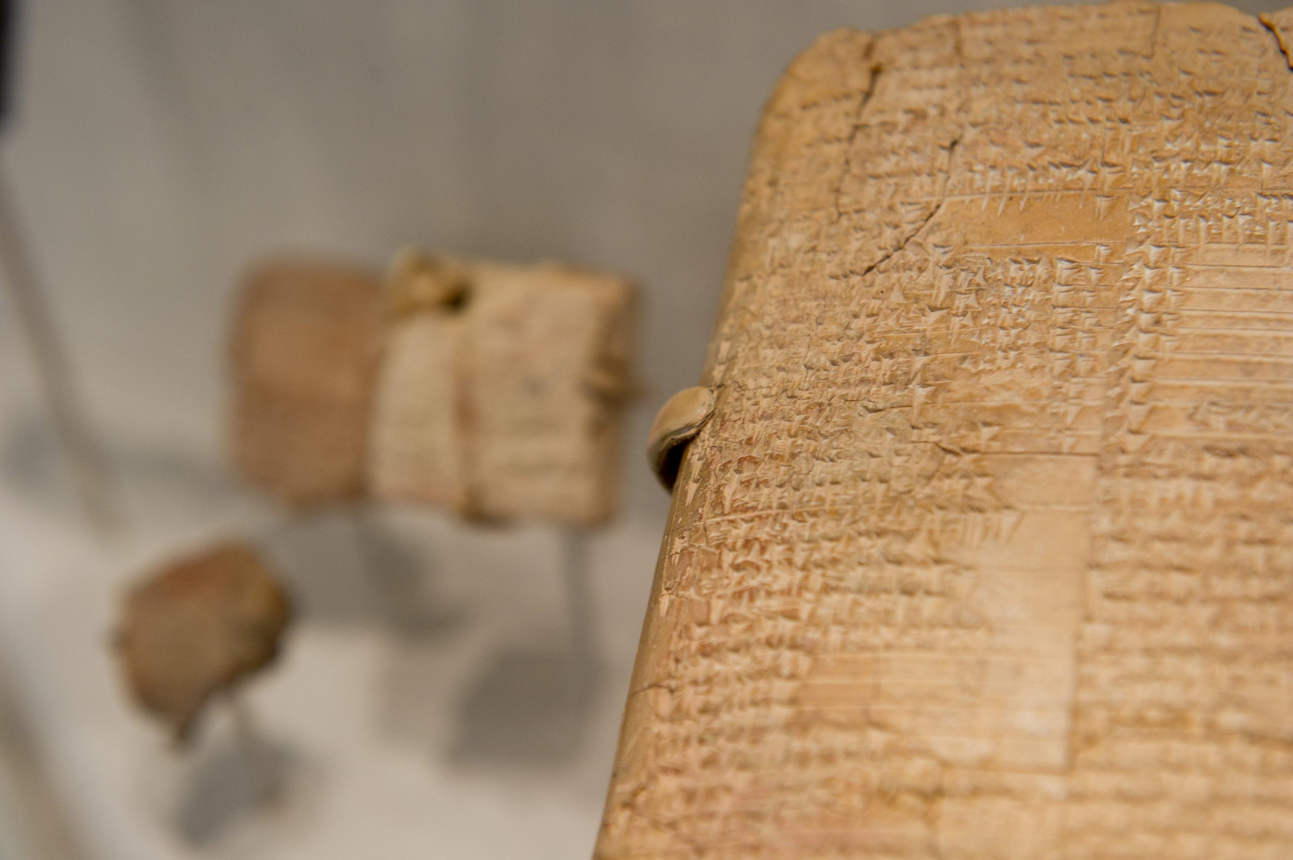 Close-up of ancient clay tablets with cuneiform script, featuring intricate carved symbols. Background shows blurred, smaller tablets on stands.