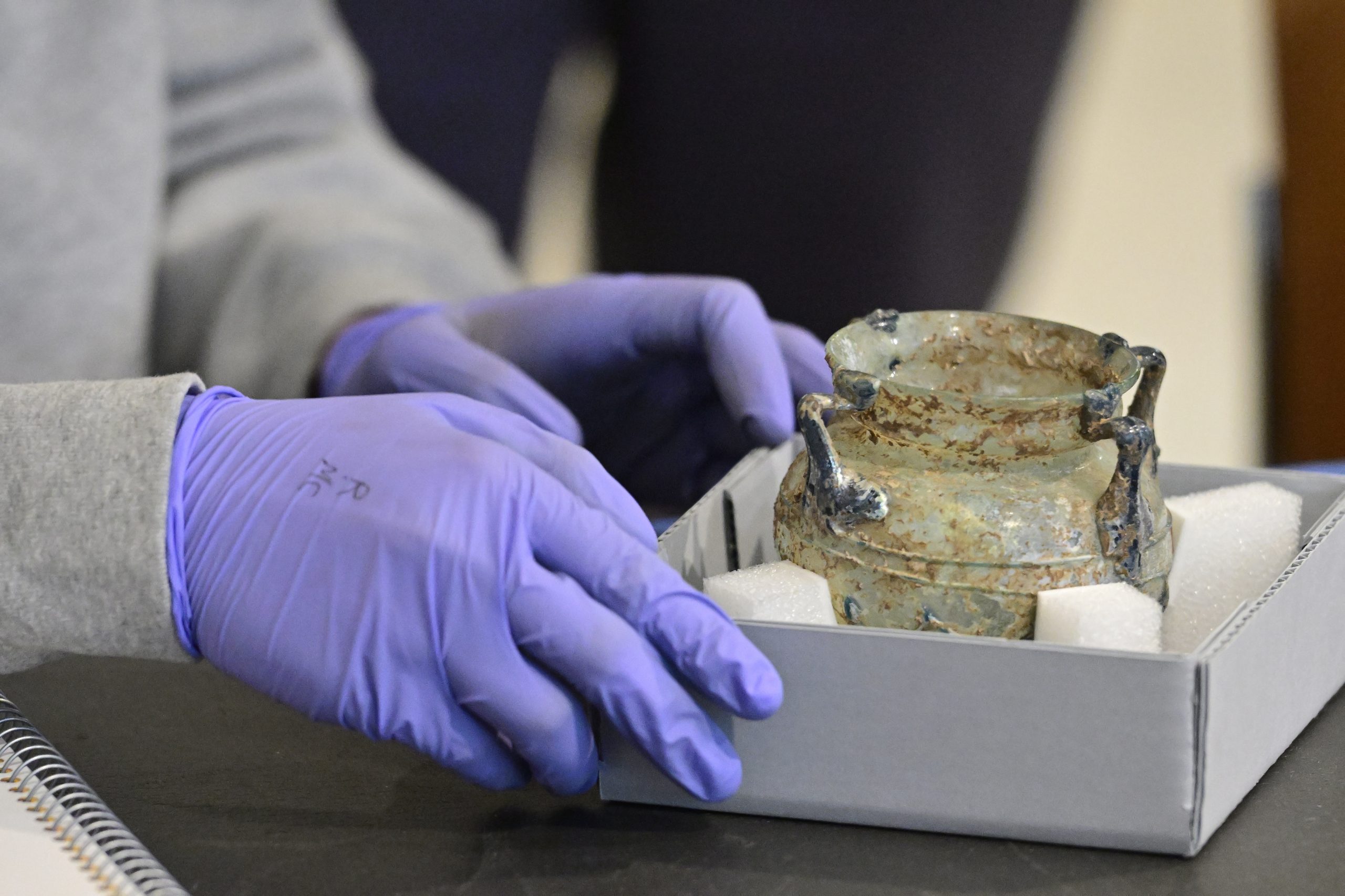 Closeup of a person wearing purple nitrile gloves holding a blue box with an ancient glass vessel.